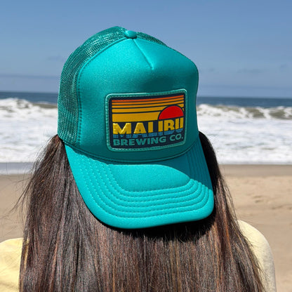 Waves and blue sky frame a person with long brown hair, wearing the Otto Sunset Patch Hat—a teal summer trucker cap featuring a colorful MAUI BREWING CO. patch—creating a stylish beach look.