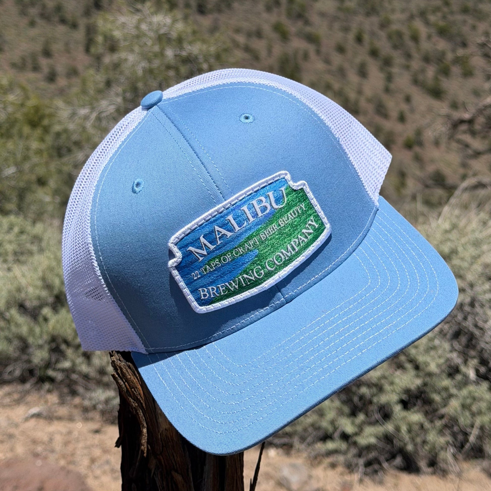 A Malibu City Sign Hat in light blue and white with an embroidered patch sits on a tree branch outdoors, surrounded by dry brush and softly blurred hills in the background.