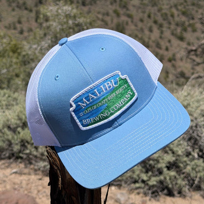 A Malibu City Sign Hat in light blue and white with an embroidered patch sits on a tree branch outdoors, surrounded by dry brush and softly blurred hills in the background.