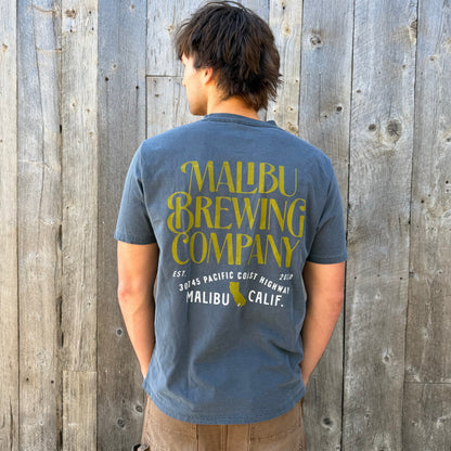 Person wearing a blue t-shirt with 'Malibu Brewing Company' text in front of a wooden wall.