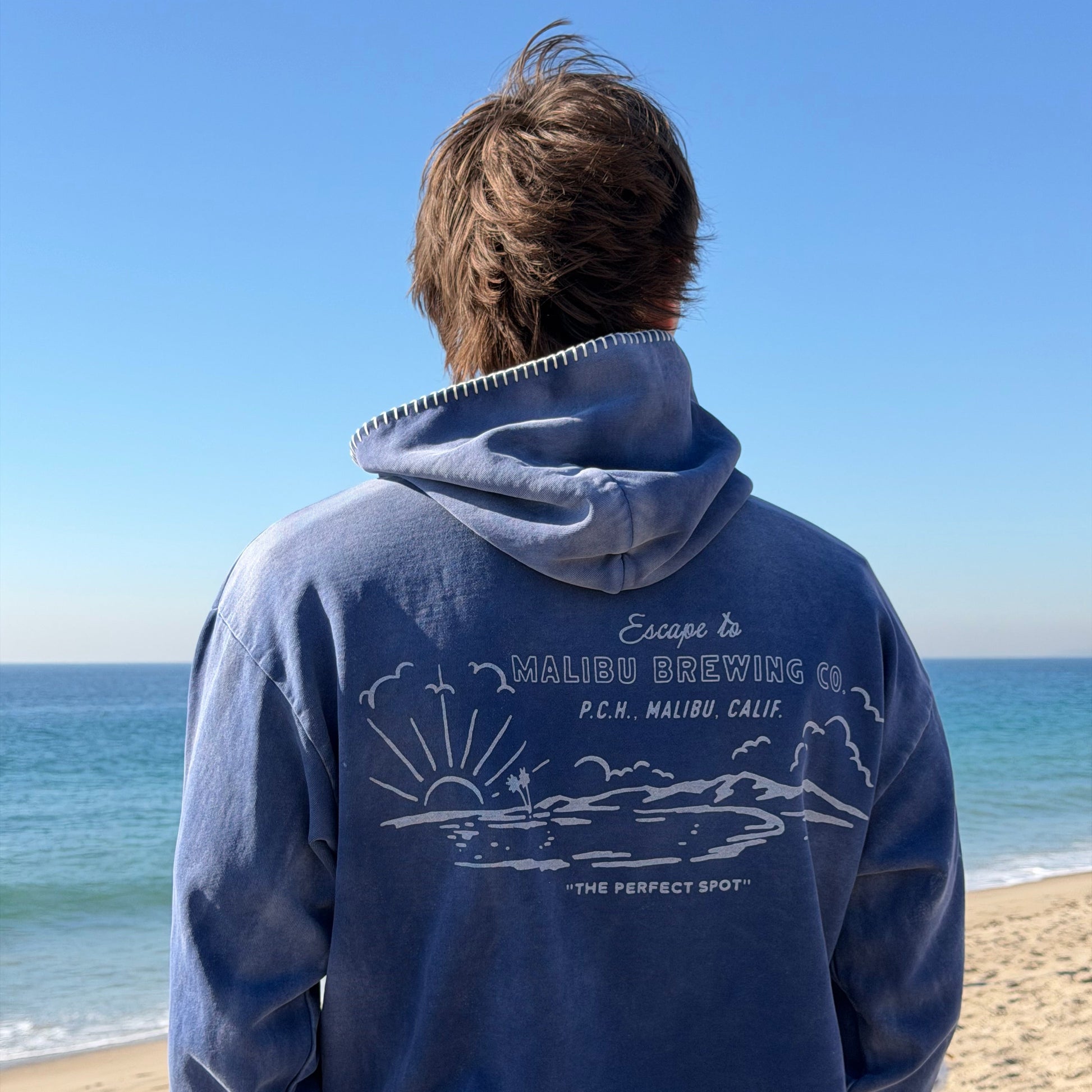 Person wearing a blue hoodie with graphics and text, standing on a beach.