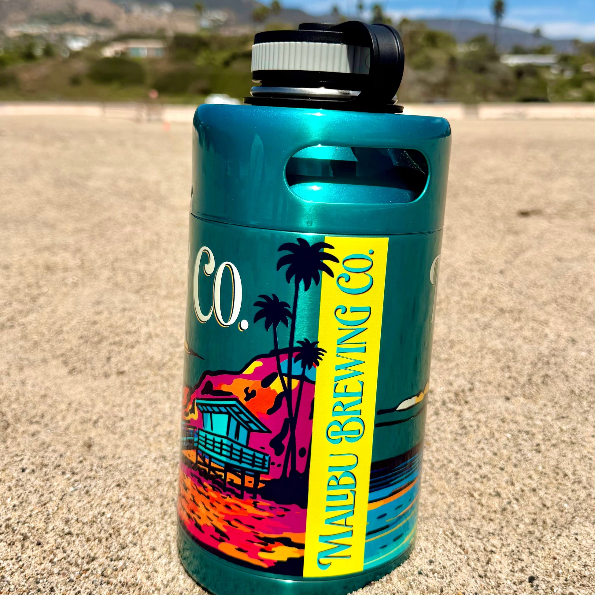 A teal Keg Growler featuring a Malibu Brewing Co. label rests on sandy ground beneath a blue sky, with palm trees and mountains in the distance—perfectly embodying California’s relaxed atmosphere. Styled like stainless growler keg growlers, this vacuum insulated Keg Growler keeps your brew refreshingly cool, even during the hottest beach days.