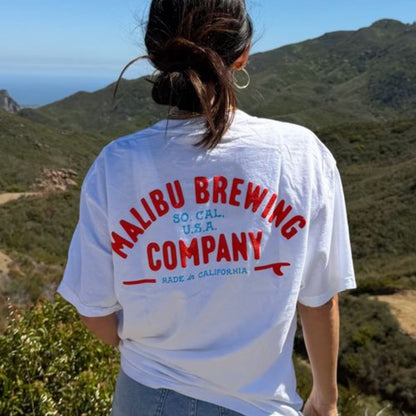 A person with long hair in a ponytail stands outdoors facing away, wearing the Red, White & 'Bu Tee from Malibu Brewing Company. Green hills and the ocean extend under a clear summer sky.