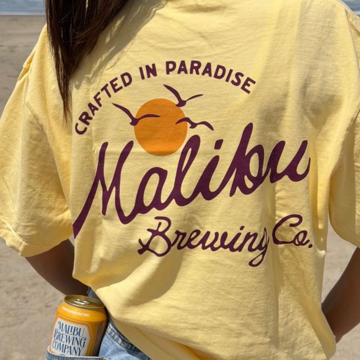 Wearing a yellow Crafted in Paradise Tee featuring a sunset, birds, and “Crafted in Paradise,” someone stands on the beach with a Malibu Brewing Co. canned drink in their back pocket, showing off laid-back coastal style.